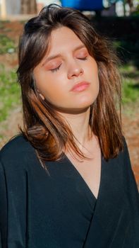 A woman with eyes closed enjoying the sunlight in an outdoor setting wearing a black blouse.